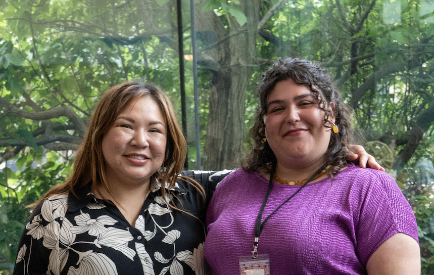 Two women are smiling with their arms around each other, visible from the waist up. 
